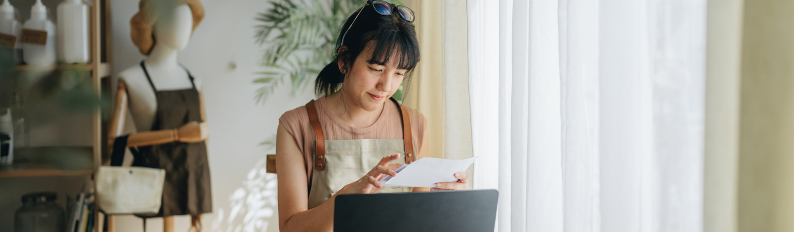 Female presenting store owner works at laptop, wearing apron.