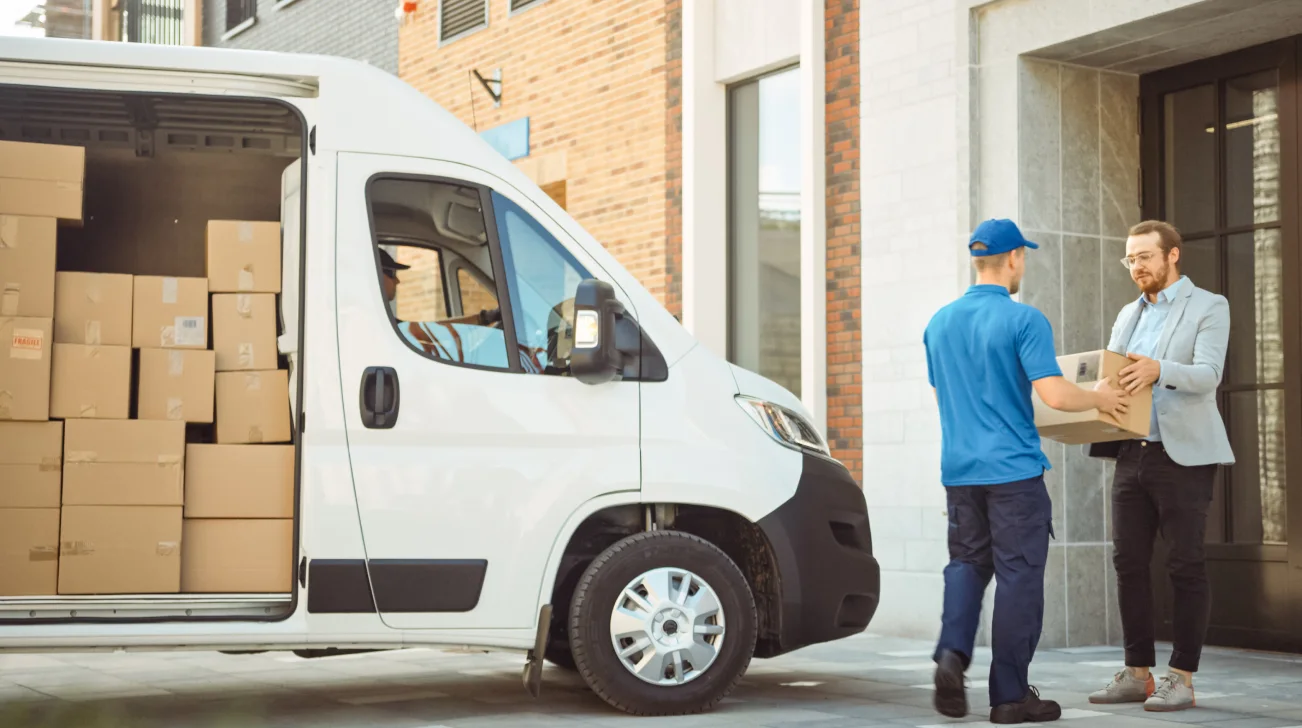 Delivery man in blue uniform handing box to customer next to delivery van containing more boxes