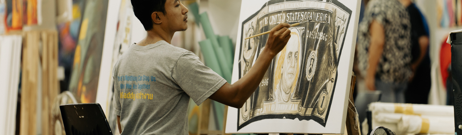 Male presenting person sits at an easel, painting an image of an American bank note.