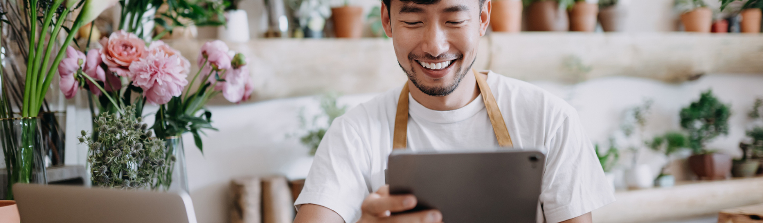 Male presenting person smiles at their ipad, surrounded by colourful plants and flowers in a shop.