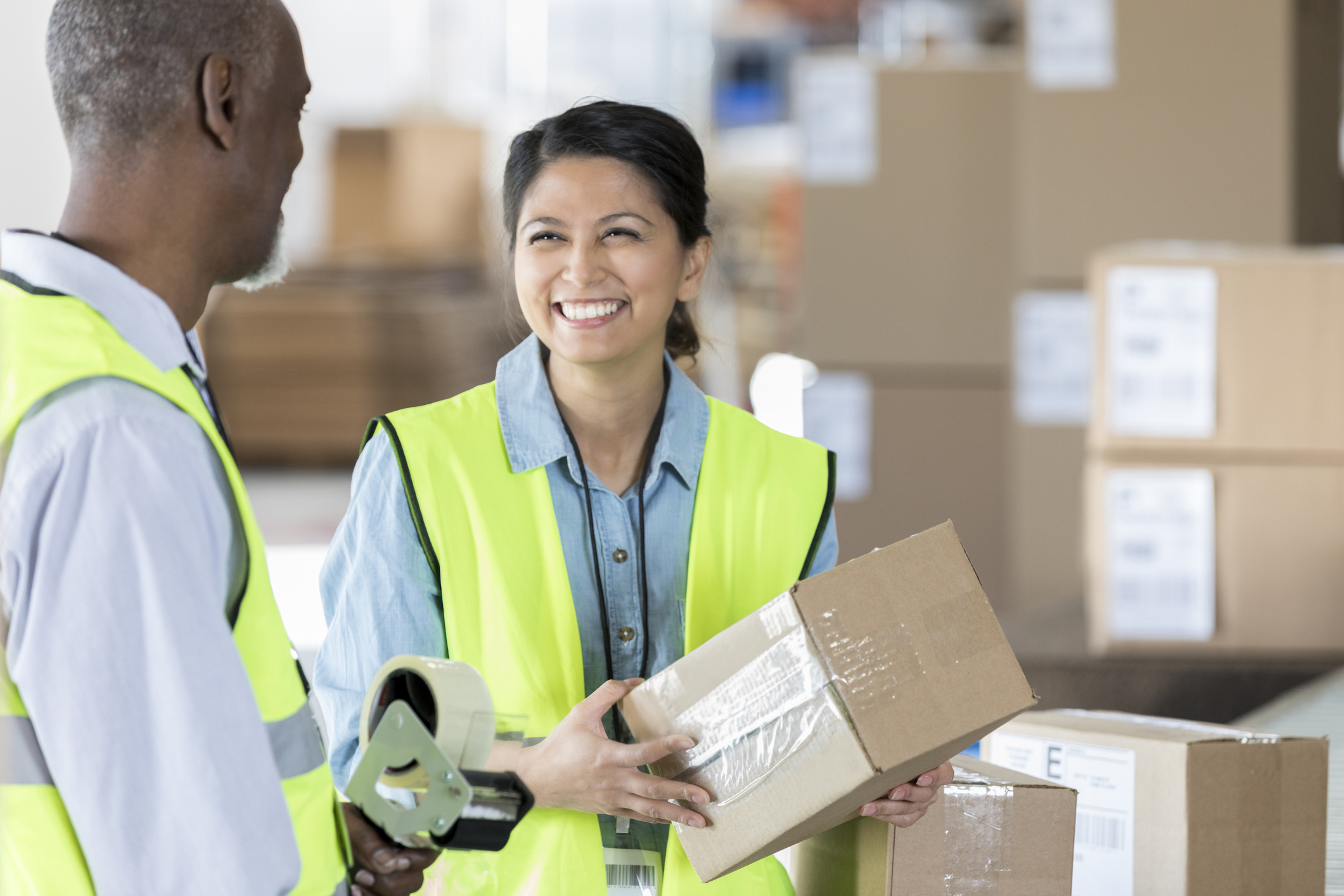 Two warehouse workers in high-visibility vests smiling and collaborating while handling a cardboard box and packing supplies, surrounded by stacked boxes in a distribution center.