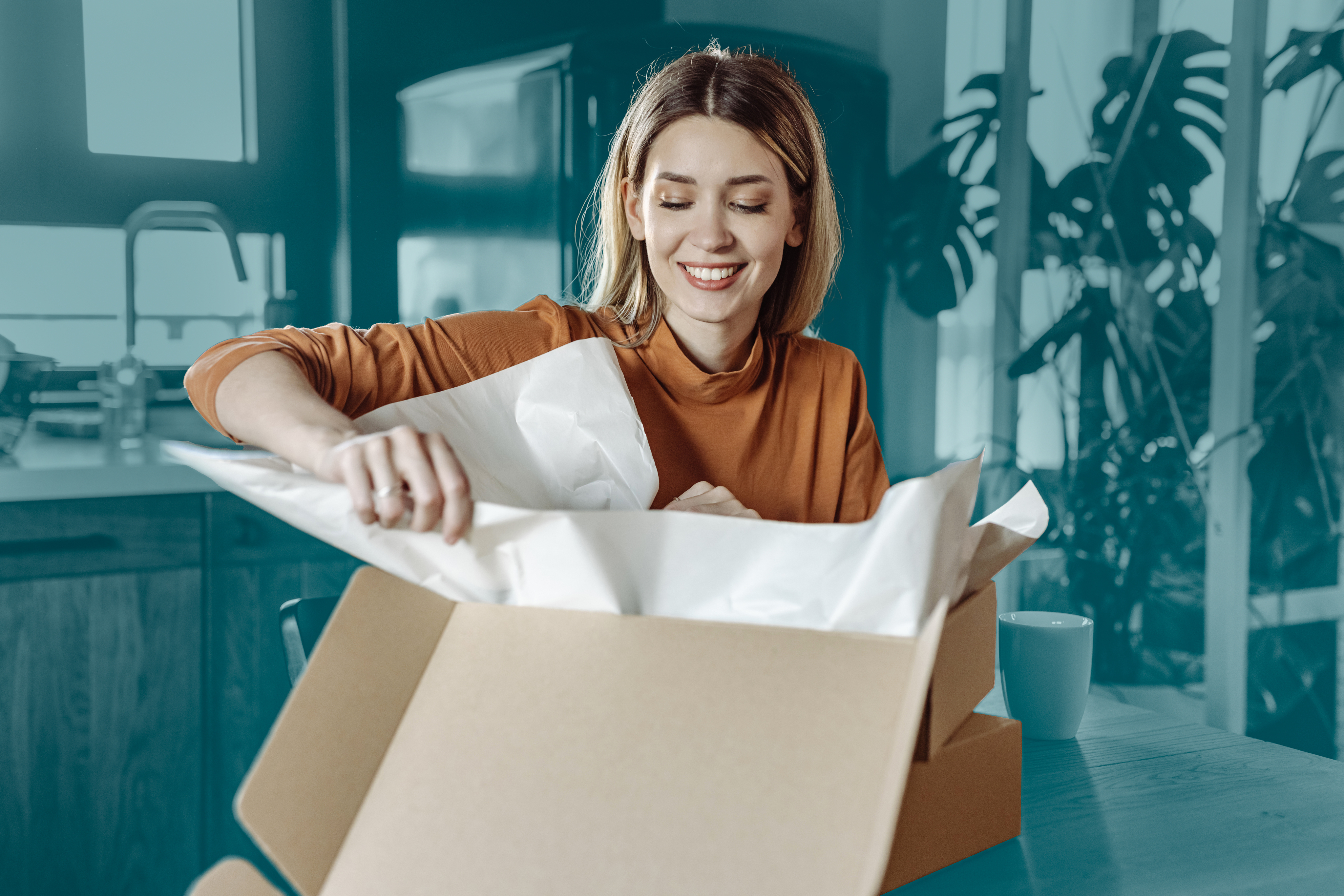 Smiling woman sitting at a table in a modern kitchen, opening a cardboard shipping box filled with white packing paper, with a coffee mug and houseplants visible in the background.
