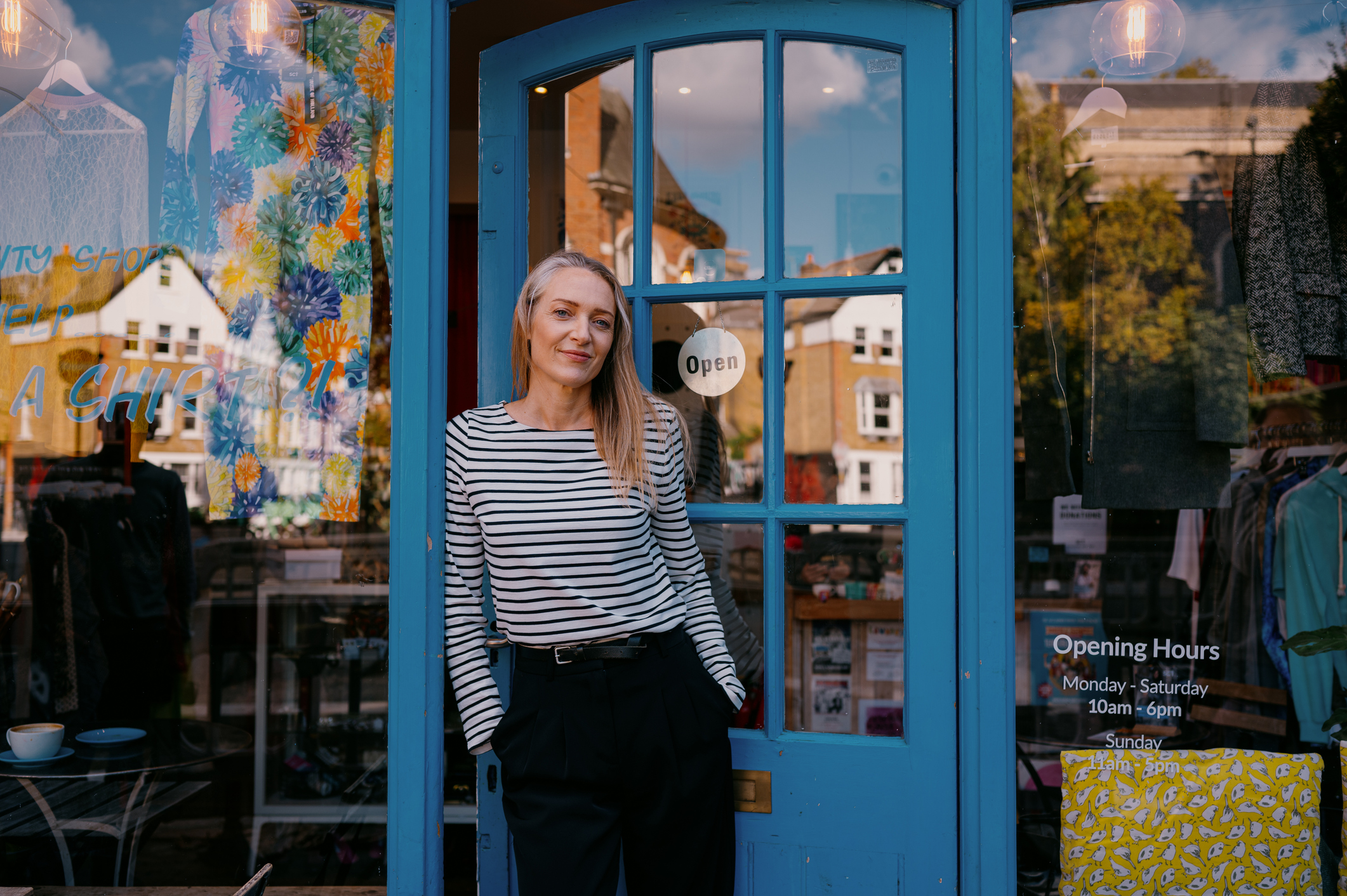 Female presenting person stands outside their shop, leaning against a blue door.