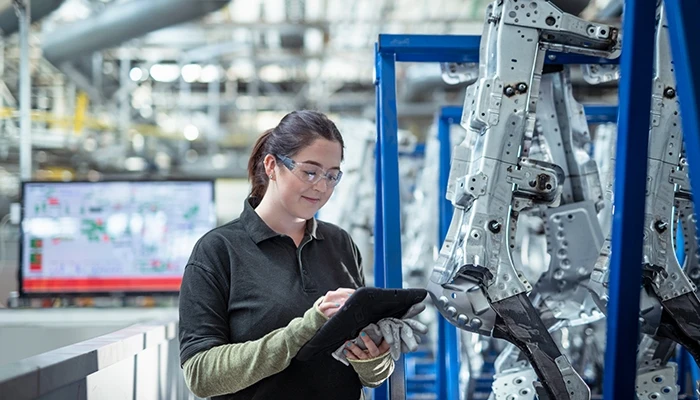 A woman in safety glasses holds a tablet on a factory floor.