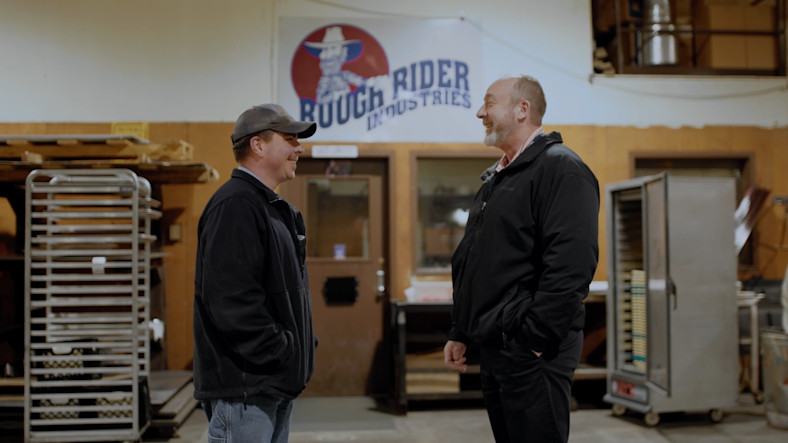 David Sisson and Rick Gardner stand in front of a banner for Rough Rider Industries
