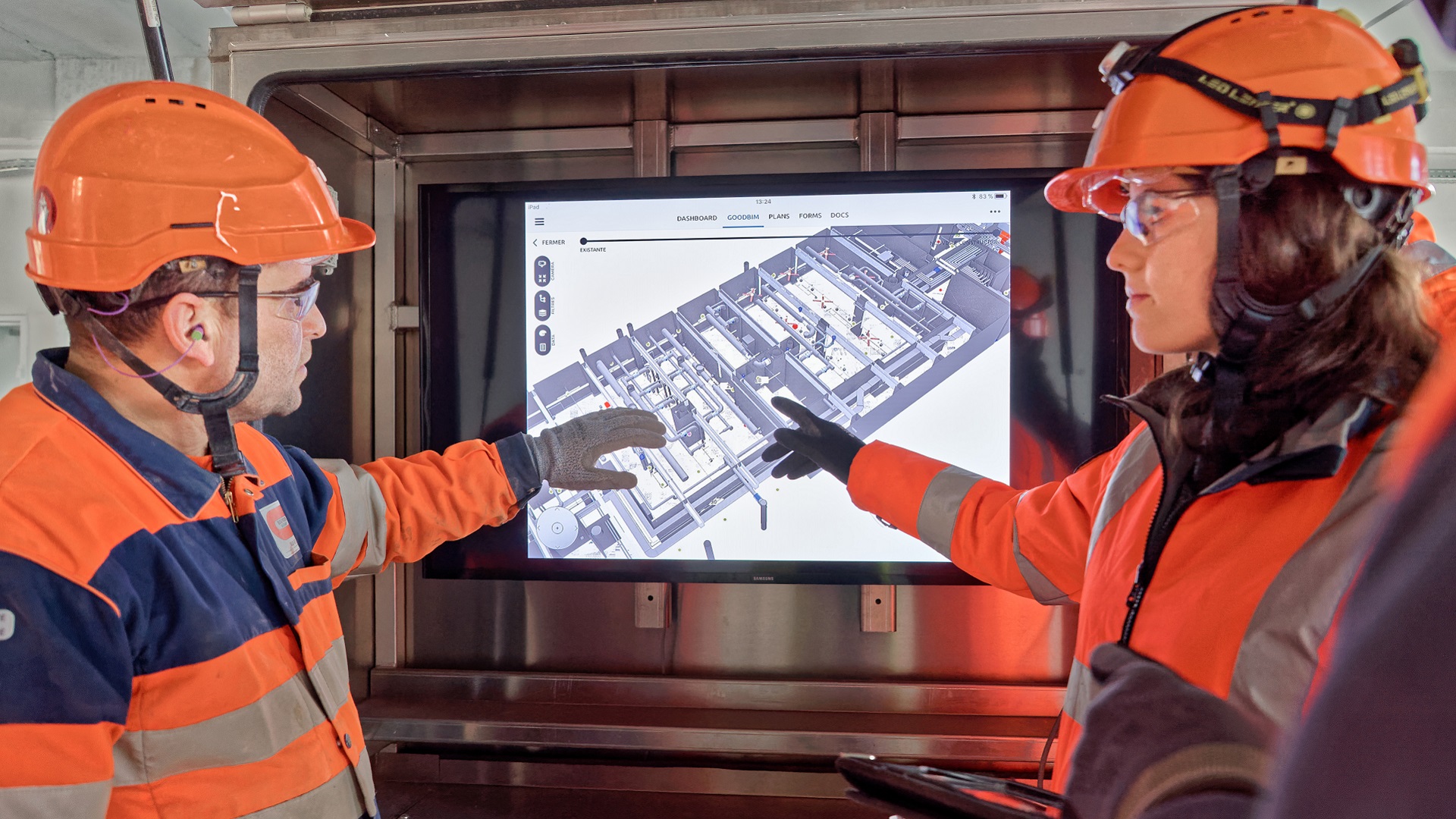 Bouygues Construction workers in hard hats point to 3D model on a monitor on a construction site.
