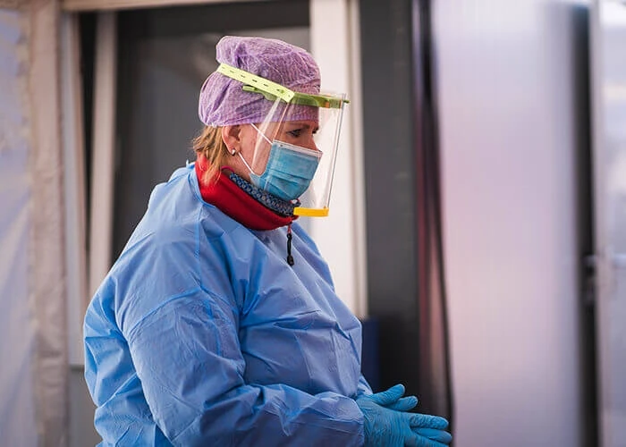 A health care worker wears PPE including surgical gown, gloves, mask, and a plastic face shield.
