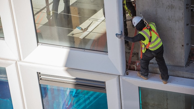 Construction workers install a large prefabricated piece containing a window and 3D-printed concrete mold on a building façade.