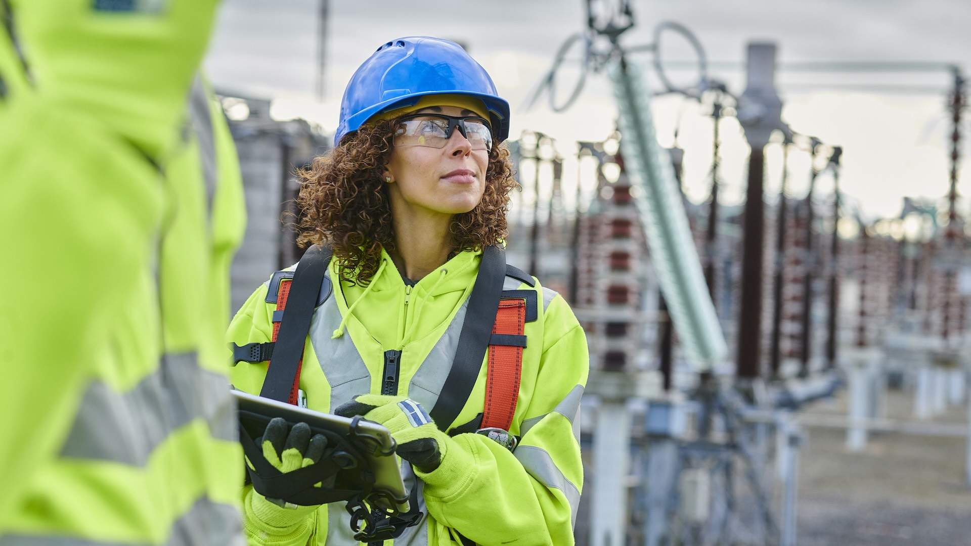 A woman in safety gear stands at a power substation holding a tablet.