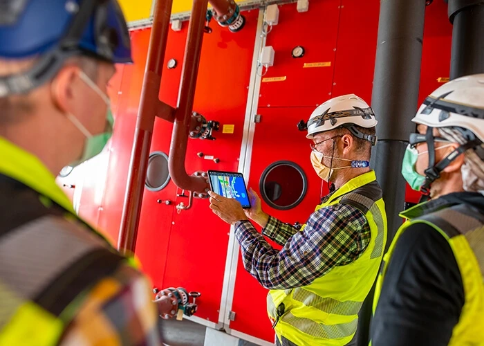 Three people in work vests, masks, and hard hats look at a tablet.