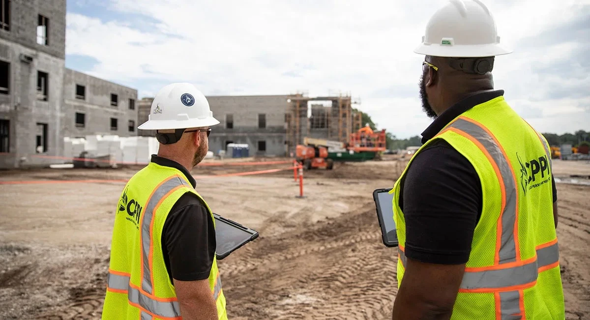 Two construction supervisors in safety vests and hard hats review an active job site with unfinished concrete buildings and equipment.