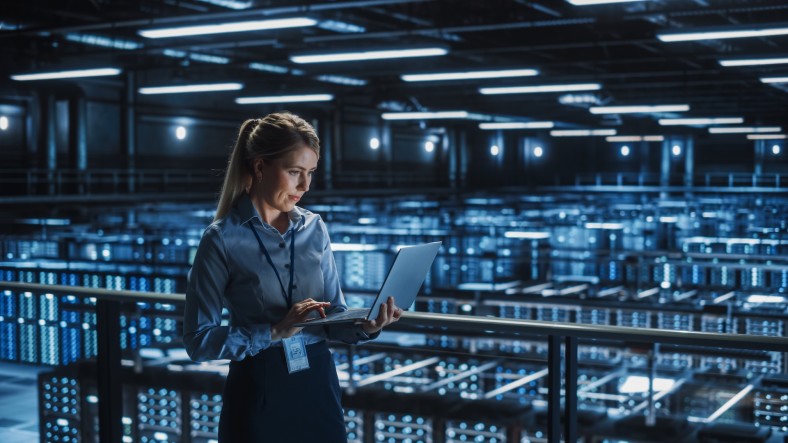 A woman with a laptop stands in a large room full of hundreds of data servers.