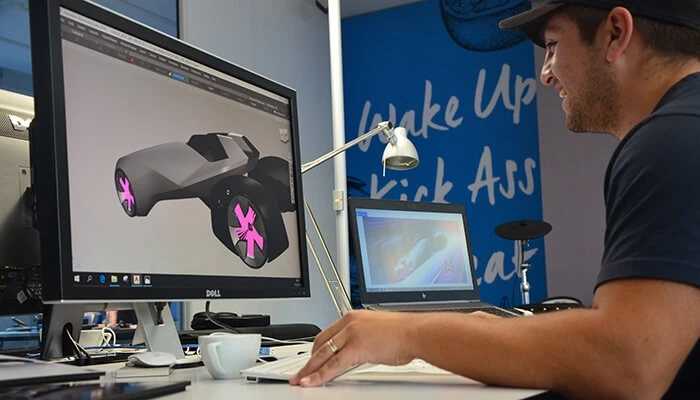 A smiling designer works on a digital sports car at a desk with two monitors.