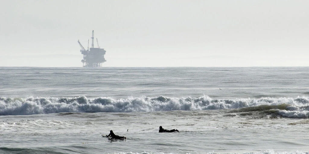 Surfers paddle out into the ocean, with an oil rig in the distance