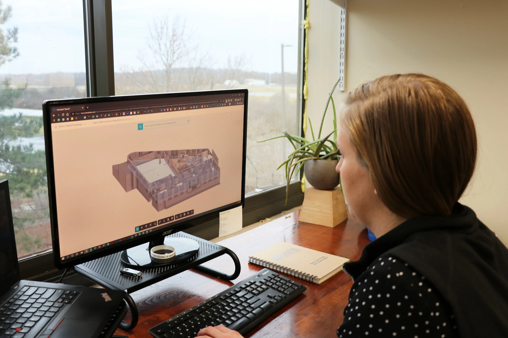 Person at a desk reviews a 3D building model on a computer monitor in an office with a window and plant nearby.