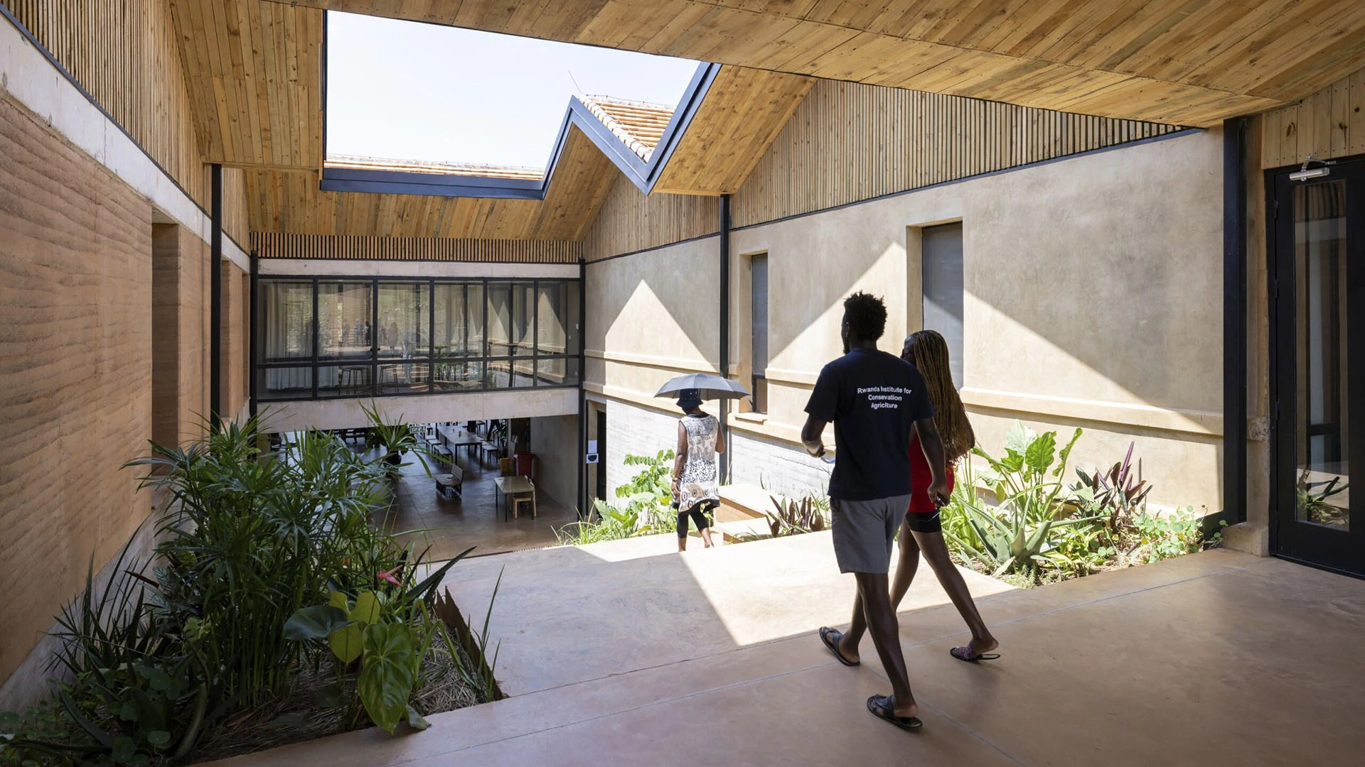 Un homme et une femme traversent une cour intérieure baignée de soleil et habillée de panneaux de bois.