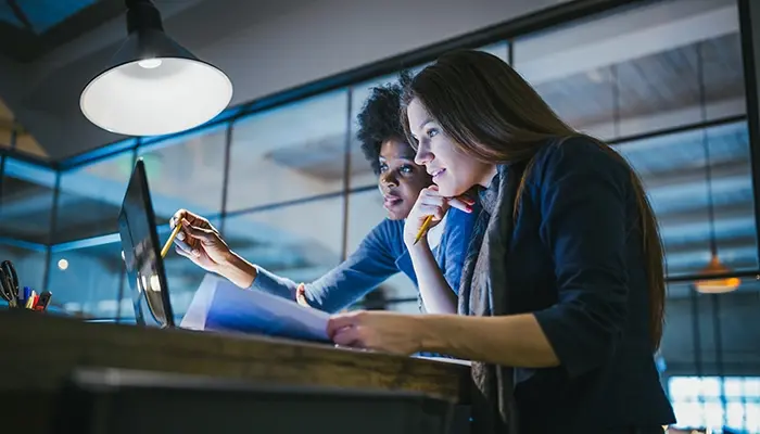 Two women in an office look at a laptop screen.