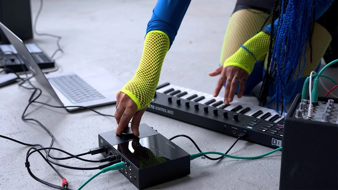 A musician adjusts a knob on a signal processing unit with one hand as they use the other to press a key on a synthesizer keyboard 
