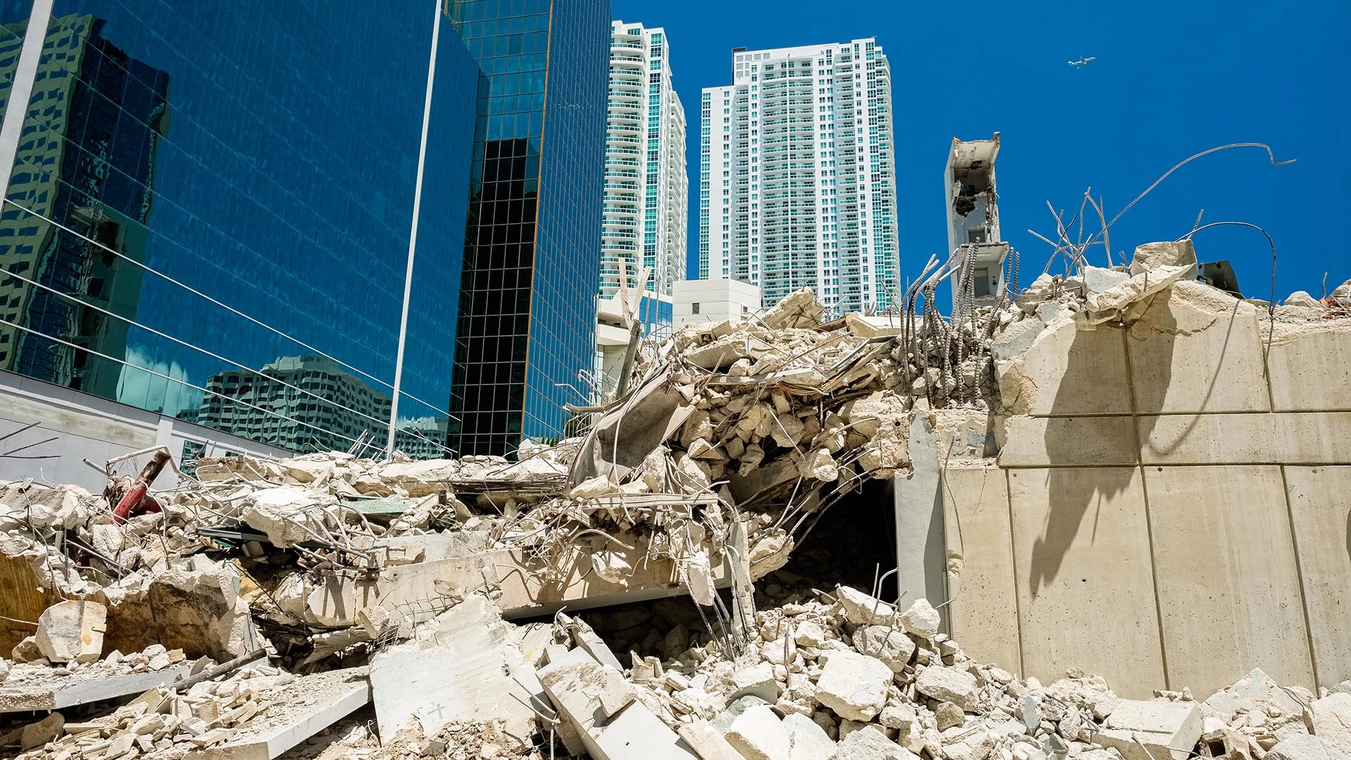 Un edificio de hormigón parcialmente demolido se alza con varillas de refuerzo expuestas y escombros entre modernos rascacielos acristalados en un entorno urbano.