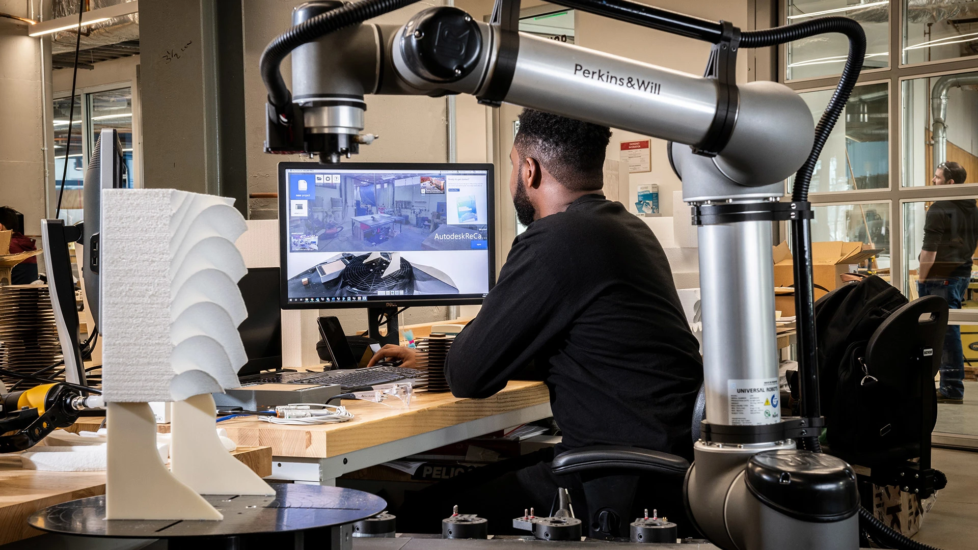 A man at a workstation is using a robot to help form a piece of architectural material.