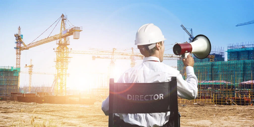 Person sitting in a director's chair wearing a hardhat and holding a megaphone, shouting at a construction site