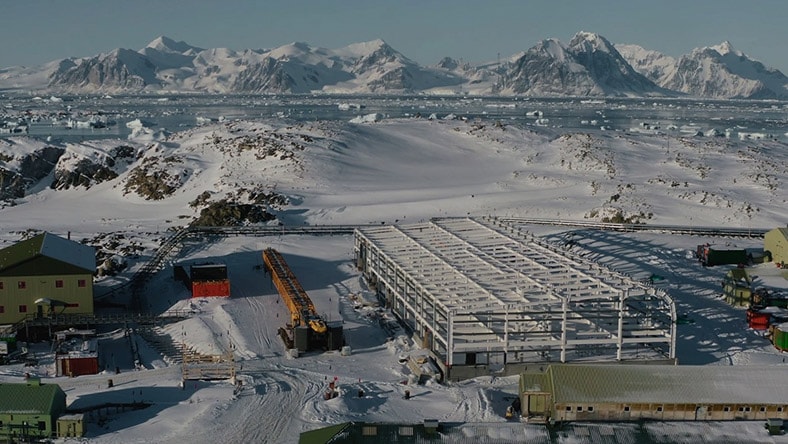 An aerial view shows facilities under construction in Antarctica, with icy waters and snow-covered mountains in the background.