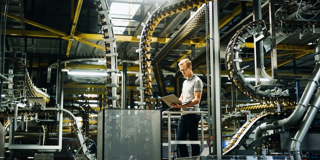 A man in glasses stands looking at laptop in a factory.