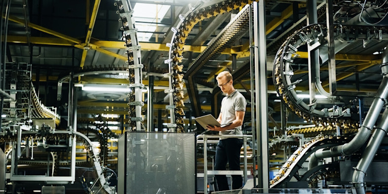 A man in glasses stands looking at laptop in a factory.