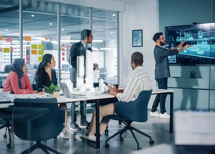 Four office workers sitting at a table collaborating while another colleague runs a presentation on a screen.