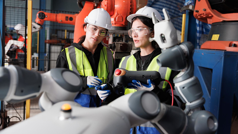 Two women in hard hats consult a control panel in a robot development plant.