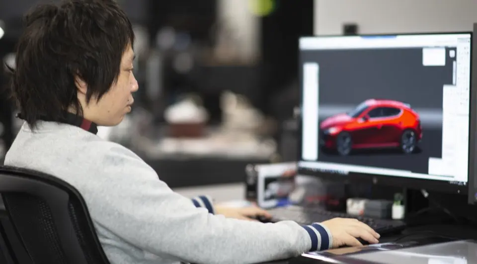 A person working on a computer, designing a red car model displayed on the monitor in a professional workspace.