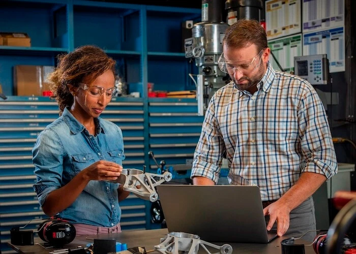 A woman in safety glasses handles a 3D-printed part next to a man in safety glasses working on a laptop.