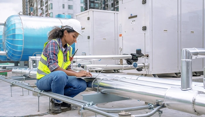 A woman in a hard hat and safety vest works on a laptop at a water treatment plant.