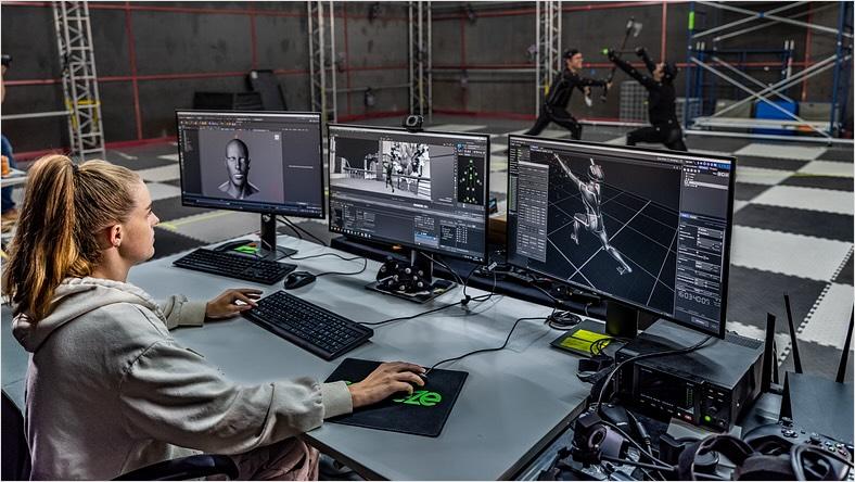 In a motion capture studio, a woman sits in front of computer monitors viewing the footage of two performers stage-fighting nearby.