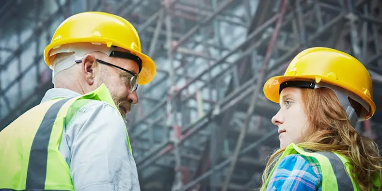 Two engineers, one middle-aged, one young, wearing vests and hard hats on a construction site
