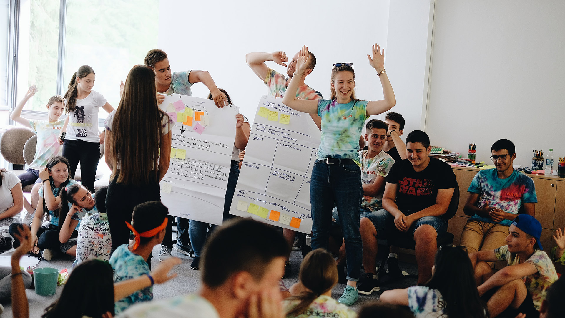 In a room crowded with teenagers, one girl stands with her arms held high, next to a poster outlining project details.