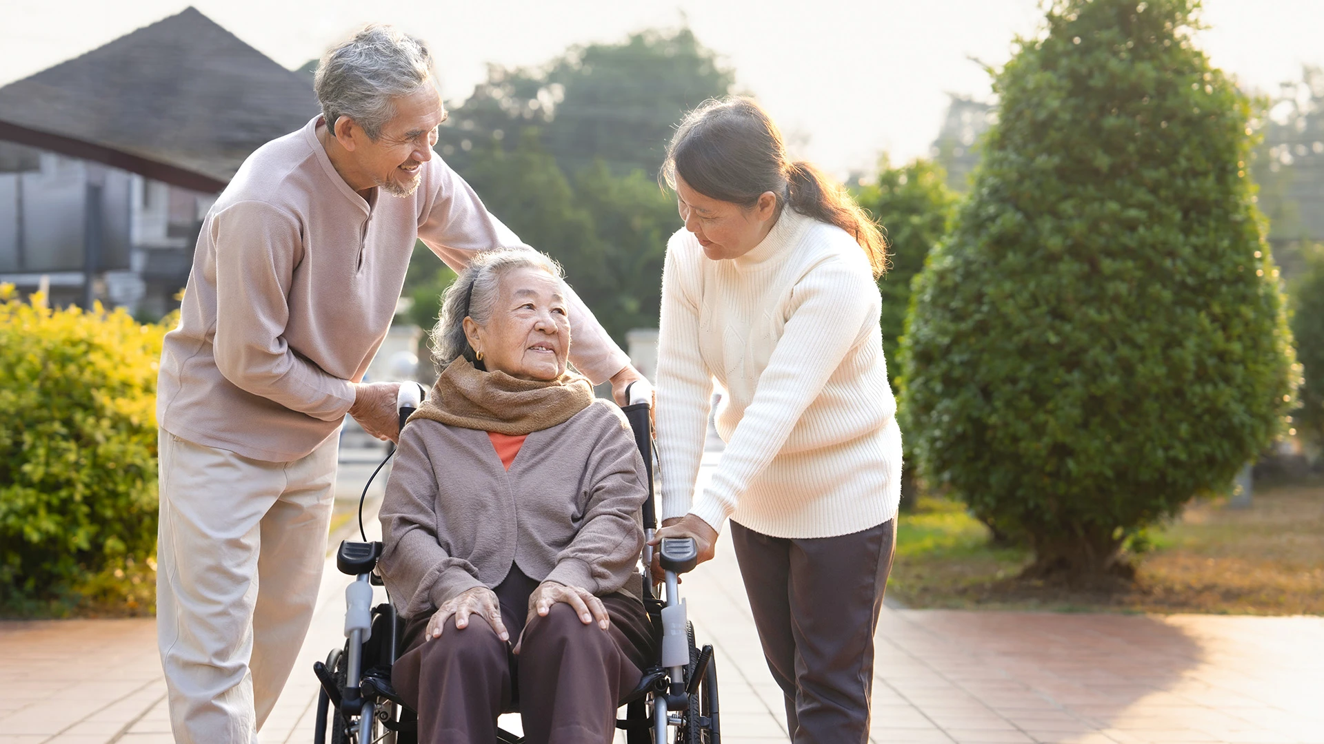 Una señora mayor, en silla de ruedas, pasea con su hijo y su nuera por el sendero de un jardín.