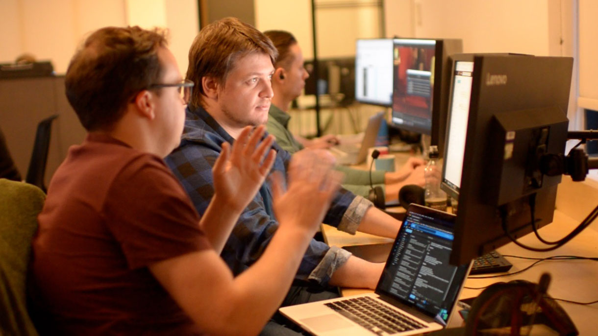Two white males collaborate in front of dual monitors and a laptop in an open-office setting