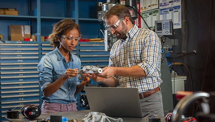 A woman and a man in safety glasses look at a metal part in a factory setting.