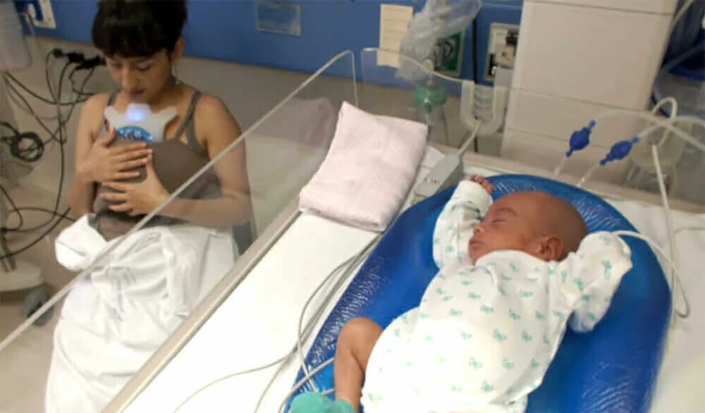 A mother sits next to her baby in a Babybe in a hospital room.