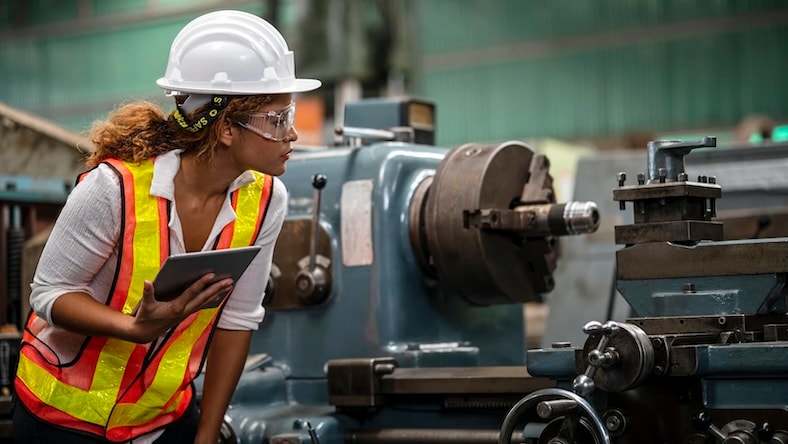 A woman in a hard hat inspects machinery while consulting a tablet.