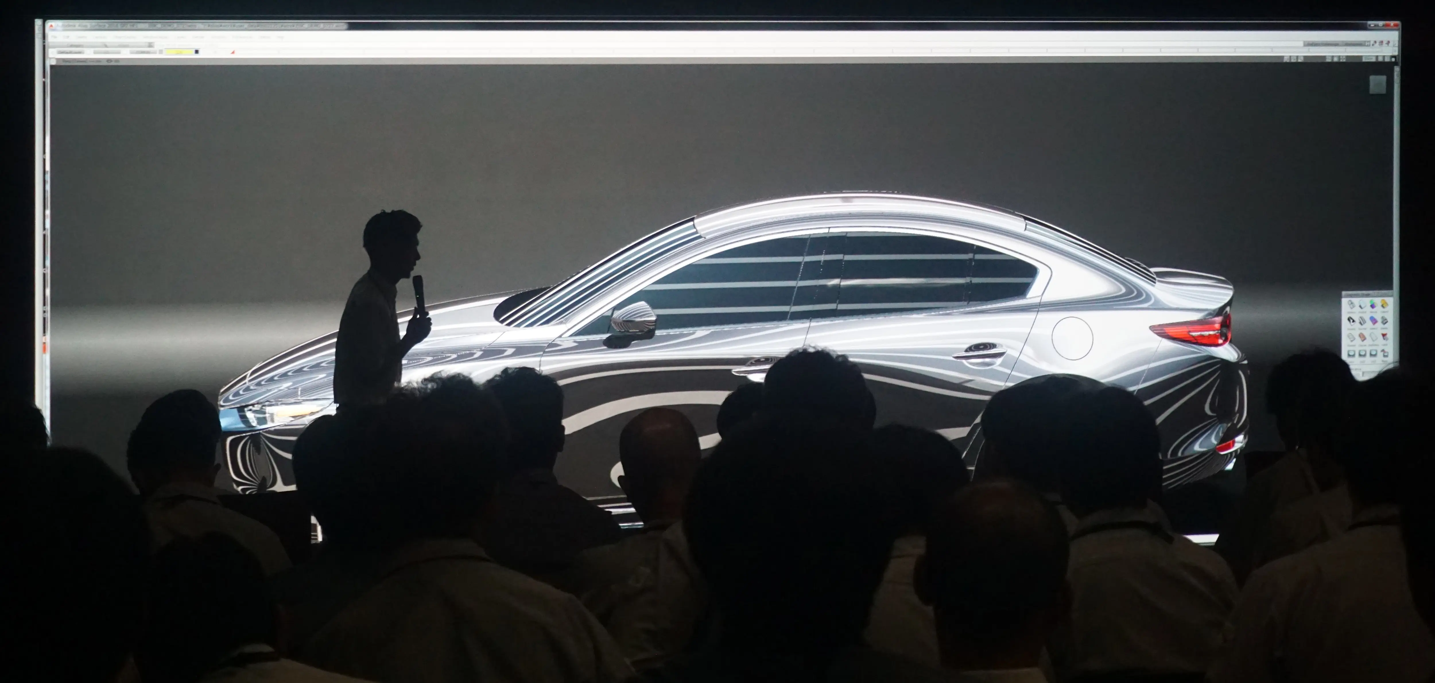 A presenter discusses a sleek car design displayed on a large screen to an audience in a dark room.