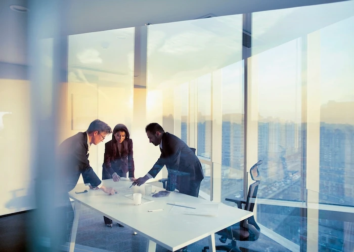 Two men and a woman stand around a desk in an office with floor-to-ceiling windows.