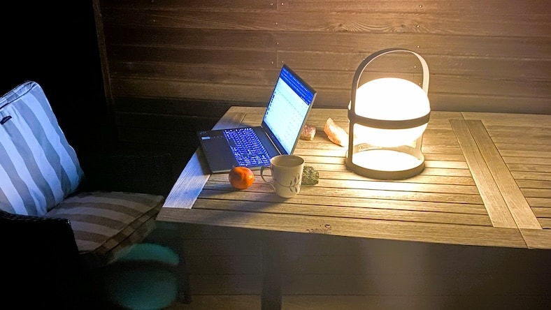 A home office set up on an outdoor table after dark includes a laptop, mug, apple, deck chair, and round, glowing portable light.