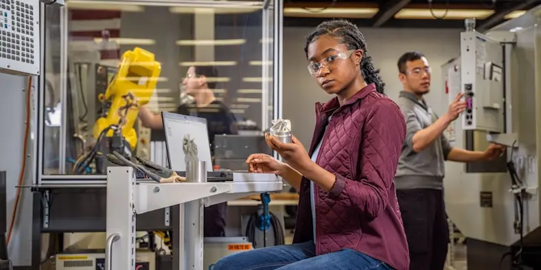 Person in a workshop using a computer and holding a product, while wearing safety goggles