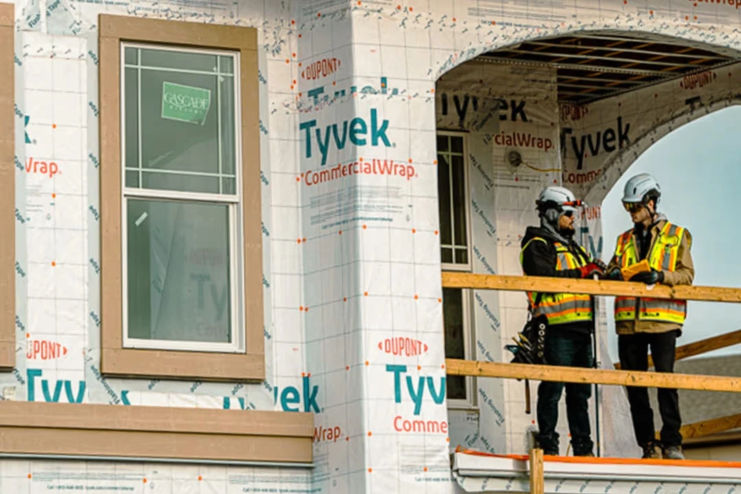 Two construction workers in safety vests stand on a balcony of a building under construction wrapped in sheathing.