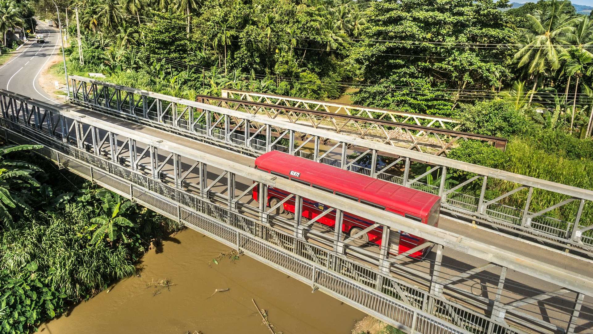 A bus crosses a steel-truss bridge over a river in a tropical landscape