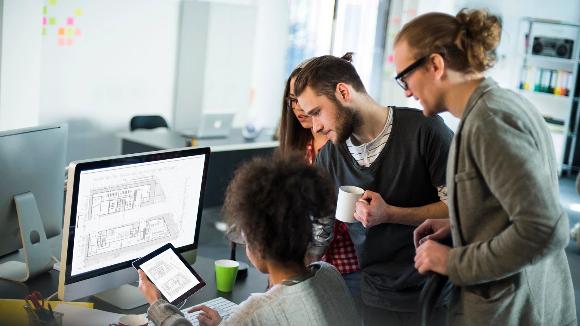 Un groupe de designers rassemblé autour d'un bureau, examinant des plans de construction numériques sur un écran et une tablette.