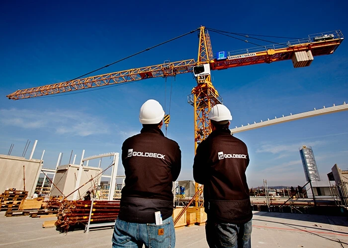 Two men in Goldbeck jackets and white hard hats stand in front of a crane