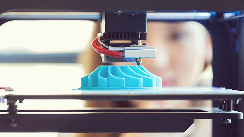 A young woman watches a printout on a 3D printer.